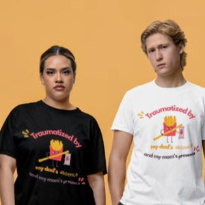 May include: A black t-shirt and a white t-shirt, each featuring a graphic of a cartoon french fry container holding a fry and a ketchup packet, with the text "Traumatized by my dad's absence and my mom's presence" printed in red and black.