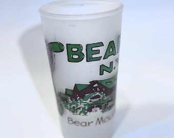 May include: A frosted glass shot glass featuring a green graphic of a building with the text BEAR MOUNTAIN N.Y. printed in black and green. The cylindrical vessel has a smooth, translucent finish and a clear rim at the top edge.
