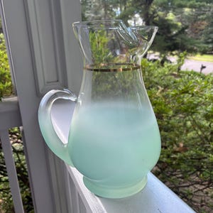 May include: A glass pitcher featuring a frosted mint green base that transitions into clear glass at the top. The pitcher includes a curved handle and a thin, metallic gold band encircling the neck, just below the wide, pourable spout.