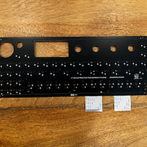 May include: A black rectangular electronic circuit board plate featuring numerous circular cutouts, a large rectangular opening, and white printed text that reads IF YOU CAN READ THIS YOUR WARRANTY IS DONE. CONGRATULATIONS. Two white ribbon cables are attached.