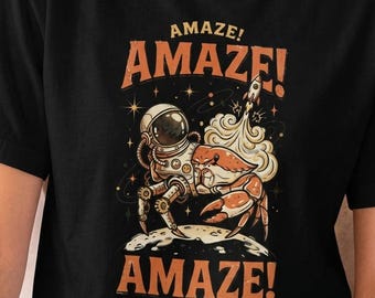 Könnte beinhalten: Ein schwarzes T-Shirt mit einem Grafikdruck eines Krebses, der einen Astronautenhelm und mechanische Beine auf einer Mondoberfläche trägt. Im Hintergrund startet eine Rakete mit Rauch. Der Text AMAZE! steht in Orange über und unter dem Motiv.