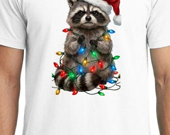 Könnte beinhalten: Ein weißes T-Shirt mit einem Grafikdruck eines Waschbären, der eine rot-weiße Weihnachtsmütze trägt. Der Waschbär ist in eine Lichterkette mit bunten roten, grünen, gelben und blauen Weihnachtslichtern verwickelt und schaut ernst.