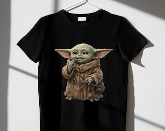 Könnte beinhalten: Ein schwarzes kurzärmeliges T-Shirt mit einem Grafikdruck einer kleinen grünen Kreatur mit großen Ohren und Augen, die eine braune Kapuzenrobe trägt und einen kleinen runden Gegenstand hält, präsentiert auf einem weißen Kleiderbügel.