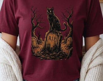 Könnte beinhalten: Ein kastanienbraunes Kurzarm-T-Shirt mit einem Grafikdruck einer schwarzen Katze, die auf einem Grabstein mit der Aufschrift RIP sitzt. Das Design zeigt kahle, dunkle Bäume und eine Friedhofskulisse mit weiteren kleineren Grabsteinen.