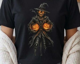 Könnte beinhalten: Ein schwarzes T-Shirt mit einem Grafikdruck einer Vogelscheuche in einem zerlumpten Mantel und mit einem breitkrempigen Hut. Die Vogelscheuche hat leuchtend orangefarbene Augen und hält zwei leuchtende Kürbisse, mit einer schwarzen Krähe auf der Schulter.