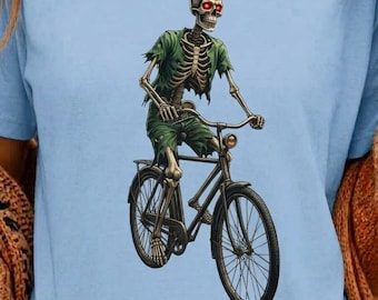 Könnte beinhalten: Ein hellblaues T-Shirt mit einem Grafikdruck eines Skeletts mit leuchtend roten Augen, das ein zerrissenes grünes T-Shirt und eine kurze Hose trägt und auf einem schwarzen Fahrrad fährt. Das Skelett ist in einer Vorwärtsposition abgebildet.