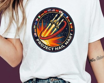 Könnte beinhalten: Ein weißes T-Shirt mit einer kreisförmigen Grafik, die drei Raketen beim Start vor einem dunkelblauen Weltraumhintergrund zeigt. Das Emblem enthält einen roten Orbitring, eine leuchtend orangefarbene Explosion und einen Rand aus internationalen Flaggen mit dem Text PROJECT HAIL MARY.