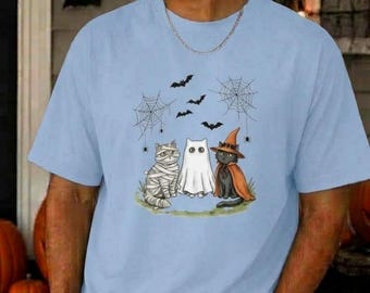 Könnte beinhalten: Ein hellblaues T-Shirt mit kurzen Ärmeln und einem Grafikdruck, der eine Mumienkatze, ein Gespenst und eine schwarze Katze mit orangefarbenem Hexenhut zeigt. Das Motiv enthält schwarze Spinnennetze, kleine Spinnen und fliegende Fledermäuse.
