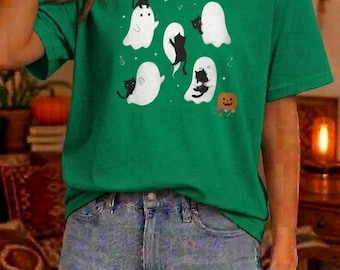 Könnte beinhalten: Ein grünes kurzärmeliges T-Shirt mit einem Grafikdruck von vier weißen Geistern, die mit kleinen schwarzen Katzen interagieren. Das Design enthält einen kleinen orangefarbenen Kürbis unten rechts, umgeben von winzigen weißen Sternen.
