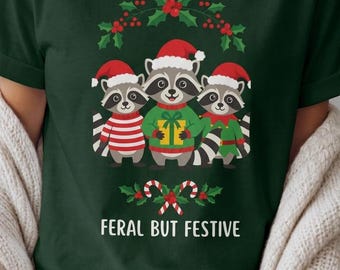 Könnte beinhalten: Ein dunkelgrünes T-Shirt mit einer Grafik von drei Waschbären, die Weihnachtsmützen tragen. Der mittlere Waschbär hält ein gelbes Geschenk. Unter den Waschbären sind Zuckerstangen und Stechpalmen sowie der Text FERAL BUT FESTIVE in Weiß.