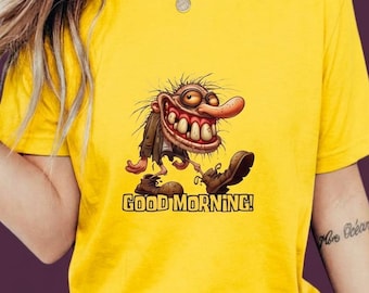 Könnte beinhalten: Ein leuchtend gelbes T-Shirt mit kurzen Ärmeln und einer grafischen Illustration einer Cartoon-Figur mit wildem Haar, großen Augen, hervorstehenden Zähnen und übergroßen Stiefeln. Darunter steht der Text GOOD MORNING! in fetten gelben Buchstaben.