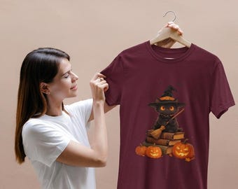 Könnte beinhalten: Ein kastanienbraunes Kurzarm-T-Shirt mit einem Grafikdruck einer schwarzen Katze, die einen Hexenhut trägt, einen Besen hält und auf einem Stapel Bücher sitzt, umgeben von drei orangefarbenen Kürbissen mit leuchtend gelben Augen und Mündern.