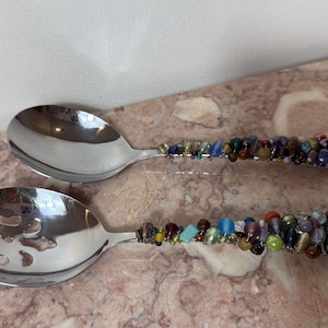 Könnte beinhalten: Ein Paar Salatbesteck aus Edelstahl, bestehend aus einem massiven Löffel und einem Schlitzlöffel. Jeder Griff ist mit einer dichten Ansammlung von mehrfarbigen Glas- und Steinperlen in Blau-, Grün-, Rot- und Brauntönen verziert.