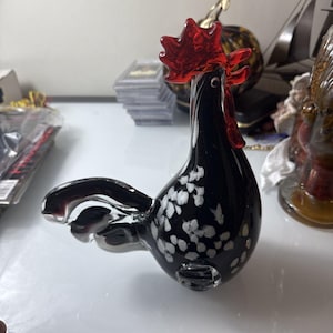 May include: A glass rooster figurine featuring a glossy black body with scattered white spots, a vibrant red comb, and a red wattle. The sculpture has a smooth, reflective surface and a curved tail, standing in a profile position.
