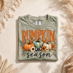 May include: A sage green crew neck t-shirt featuring a graphic print with the text PUMPKIN season in orange and black. The design includes three pumpkins in orange, teal, and white, surrounded by sunflowers, autumn leaves, and a black and white plaid bow.