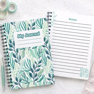 May include: A spiral-bound journal with a white cover featuring a green and teal botanical leaf pattern and the text My Journal. Beside it, an open page with horizontal lines and the word Notes. A light green pen is positioned to the left.