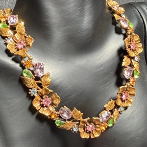May include: A gold-tone metal necklace featuring a series of interconnected floral links with textured leaves. The design is accented with faceted pink, light green, and amber-colored crystals set within the centers and between the flower motifs.