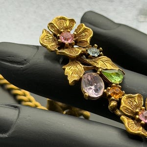 Könnte beinhalten: Ein Armband aus goldfarbenem Metall mit Blumenmotiven, die strukturierte Blütenblätter und Blätter aufweisen. Das Design ist mit facettierten, bunten Strasssteinen in Rosa, Hellblau, Limettengrün und Orange verziert, die in den Blütenmitten sitzen.