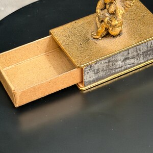 May include: A rectangular matchbox-style container featuring a gold-toned finish with a textured surface. A small, three-dimensional gold-colored cherub figure with wings is attached to the top lid. The sliding inner tray is made of plain tan cardboard.