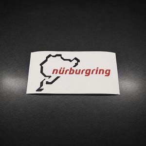 Op de afbeelding: Een rechthoekige witte sticker met een zwarte omtrek van het Nürburgring-racecircuit en de rode tekst nürburgring rechts van de circuitafbeelding. Het ontwerp is gedrukt op een witte achtergrond.