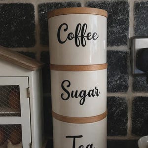 Può includere: Una pila di tre barattoli cilindrici in ceramica bianca con coperchi in legno chiaro. Ogni barattolo presenta una scritta in corsivo nero che riporta rispettivamente Coffee, Sugar e Tea. I contenitori sono disposti verticalmente.