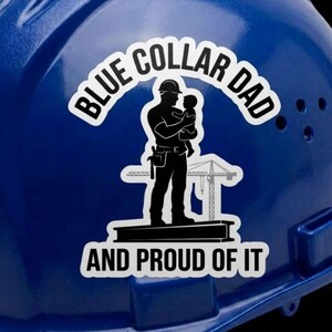 Puede incluir: Un casco de seguridad azul brillante con una pegatina blanca que muestra la silueta negra de una persona sosteniendo a un niño sobre una viga. El texto dice BLUE COLLAR DAD AND PROUD OF IT, con un gráfico de una grúa de fondo.