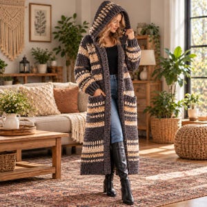 May include: A long, hooded crochet cardigan featuring horizontal stripes in dark charcoal grey and cream. The garment has long sleeves, two large front patch pockets, and an open front design worn over a black top and blue denim jeans.