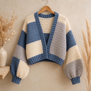 May include: A cropped crochet cardigan featuring a color-blocked pattern in shades of blue, cream, and grey. The garment has a V-neckline, long balloon sleeves with ribbed cuffs, and a ribbed hem, all constructed with a visible textured stitch.