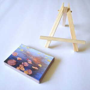 May include: A small painting on canvas depicting a blue and orange water scene with yellow and orange lily pads. The painting is displayed on a small wooden easel.
