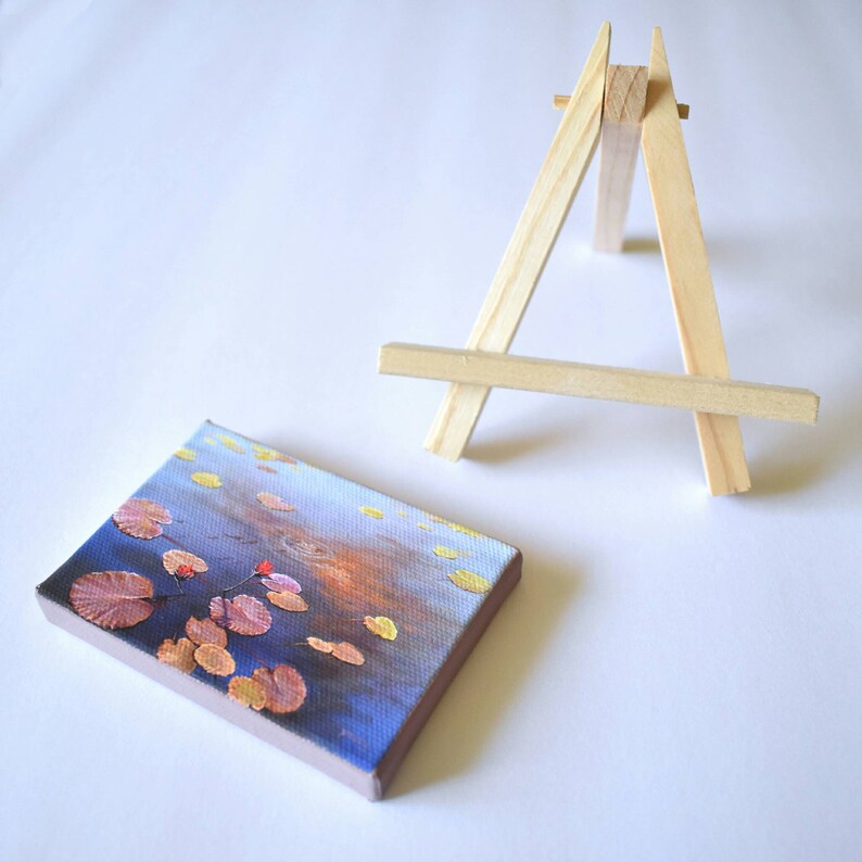 May include: A small painting on canvas depicting a blue and orange water scene with yellow and orange lily pads. The painting is displayed on a small wooden easel.