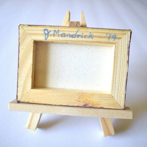 May include: A small wooden framed canvas painting on a miniature easel. The canvas is blank and the back of the frame is signed "J. Mandrick '19".