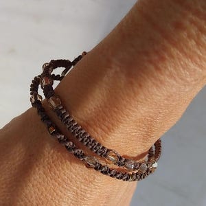 Op de afbeelding: Een dubbel gewikkelde armband van donkerbruin geknoopt koord, geaccentueerd met kleine, doorschijnende, lichtreflecterende kralen. Het koord is gevlochten in een strak, herhalend patroon en de kralen zijn gelijkmatig over de lengte verdeeld.