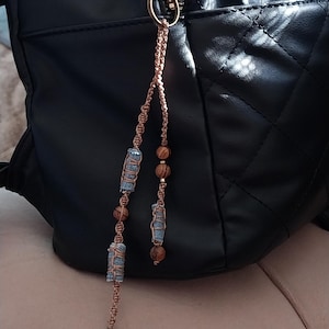 Könnte beinhalten: Ein schwarzer gesteppter Rucksack mit einem Reißverschlussanhänger aus roségoldenem Metall, an dem ein Makramee-Anhänger befestigt ist. Der Anhänger besteht aus zwei geflochtenen roségoldenen Kordeln mit zylindrischen Jeansperlen und braunen Holzperlen.