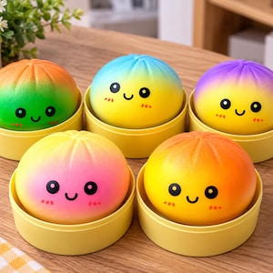 Könnte beinhalten: Fünf runde, weiche Spielzeuge in Form von gedämpften Brötchen, die jeweils in einem kleinen gelben Kunststoffbehälter liegen. Die Spielzeuge haben Farbverläufe in Orange-Grün, Blau-Gelb, Violett-Gelb, Pink-Gelb und Orange-Gelb sowie aufgedruckte schwarze Augen, lächelnde Münder und kleine rote Wangen.
