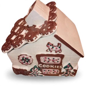 May include: A ceramic cookie jar shaped like a house with a white roof featuring brown circular decorations. The front displays a brown door with the word COOKIES, a small cat figure, a circular window, and two stylized trees with pink and green dots.