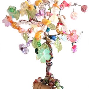 May include: A decorative wire tree sculpture featuring a cork base and branches adorned with a variety of colorful glass and plastic beads, including flower-shaped charms, translucent green leaves, and spherical beads in pink, yellow, and blue.