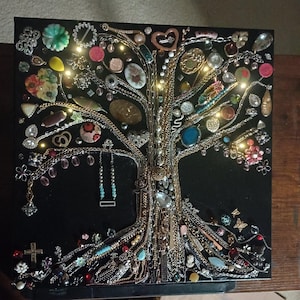 May include: A square black wall art piece featuring a tree design constructed from various vintage jewelry pieces, including gold and silver chains, colorful brooches, beads, and charms, illuminated by small embedded LED lights.