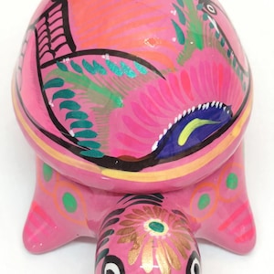May include: A pink ceramic turtle figurine featuring a hand-painted shell with vibrant floral and leaf patterns in shades of green, blue, and orange. The head is decorated with a gold flower design and black eye markings, with green accents on the legs.