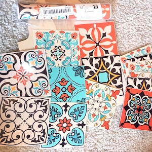 May include: A collection of square adhesive tile stickers featuring diverse decorative patterns in shades of orange, turquoise, black, and white. The set includes various floral and geometric motifs, with some stickers still contained in a clear plastic roll.