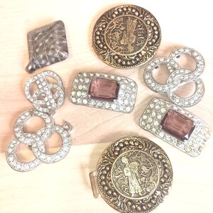 May include: A collection of seven decorative metal buckles and buttons. Included are two round brass-toned pieces with embossed figures, two rectangular silver-toned buckles with purple center stones and clear rhinestones, and three silver-toned pieces.