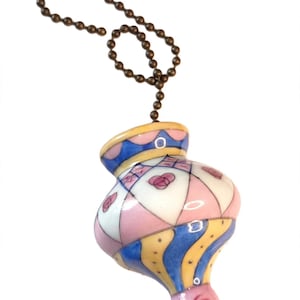 May include: A ceramic ceiling fan pull chain ornament featuring a bulbous shape with a hand-painted diamond pattern in pink, white, and blue, accented by small pink rose motifs and yellow stripes, suspended from a metallic bronze ball chain.