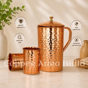 Puede incluir: Una jarra de cobre martillado con tapa y asa, acompañada de dos vasos de cobre a juego. El conjunto presenta iconos circulares con el texto BOOST IMMUNITY, IMPROVES SKIN, AIDS DIGESTION y ENHANCES METABOLISM. Copper Aura India está impreso en la parte inferior.