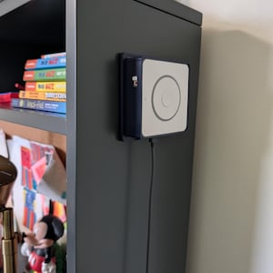 May include: A square, dark blue and white electronic device with a circular button in the center, mounted on the side of a dark gray shelf. A black braided power cable extends downward from the bottom of the device, which features a small green and red indicator light on its side.