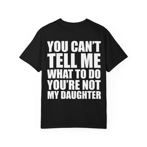 May include: A black short-sleeved t-shirt featuring bold white text printed on the back. The text reads: YOU CAN'T TELL ME WHAT TO DO YOU'RE NOT MY DAUGHTER. The garment has a crew neckline and a standard fit, displaying the message in clear capital letters.