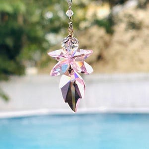 May include: A hanging suncatcher ornament featuring a faceted pink crystal shaped like a flower with a leaf-like pendant below. The crystal is attached to a silver-toned metal chain with small clear beads and a decorative metal cap at the top.