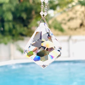 May include: A clear, multifaceted diamond-shaped crystal pendant suspended from a delicate silver-toned chain. The crystal features sharp geometric cuts that refract light, creating visible rainbow spectrums across its transparent surface.