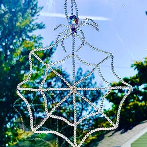May include: A hanging suncatcher ornament featuring a spider and a web constructed from clear, faceted beads. The spider has a large, iridescent crystal body and eight beaded legs, suspended by a thin metal chain against a bright outdoor view.
