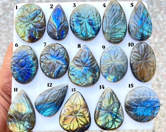 Puede incluir: Una colección de quince piedras preciosas de labradorita pulida dispuestas en una cuadrícula, numeradas del uno al quince. Estas piedras presentan intrincadas tallas florales y exhiben destellos iridiscentes de color azul, dorado y gris.