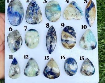May include: A collection of fifteen polished cabochon gemstones arranged on a white display card, numbered one through fifteen. Each stone features unique patterns of blue, white, cream, and metallic gold inclusions in various shapes including oval, teardrop, and marquise.