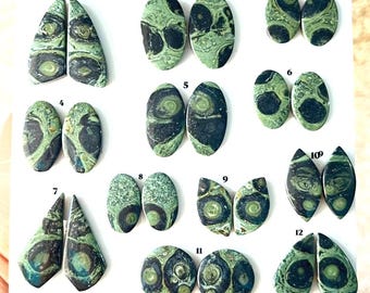 Puede incluir: Una colección de doce pares de cabujones de riolita orbicular verde y negra dispuestos sobre una superficie blanca. Cada par está numerado del uno al doce, presentando patrones circulares distintos y texturas arremolinadas.